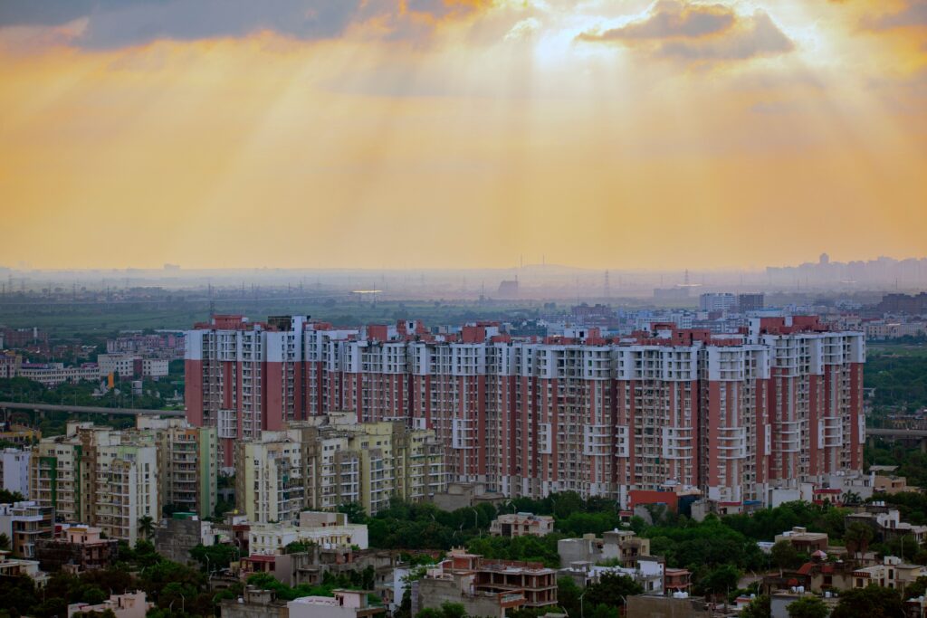 Dense row of red-and-white high-rise apartment towers rising above low-rise homes and trees, under a hazy golden sky with sunbeams breaking through clouds.
